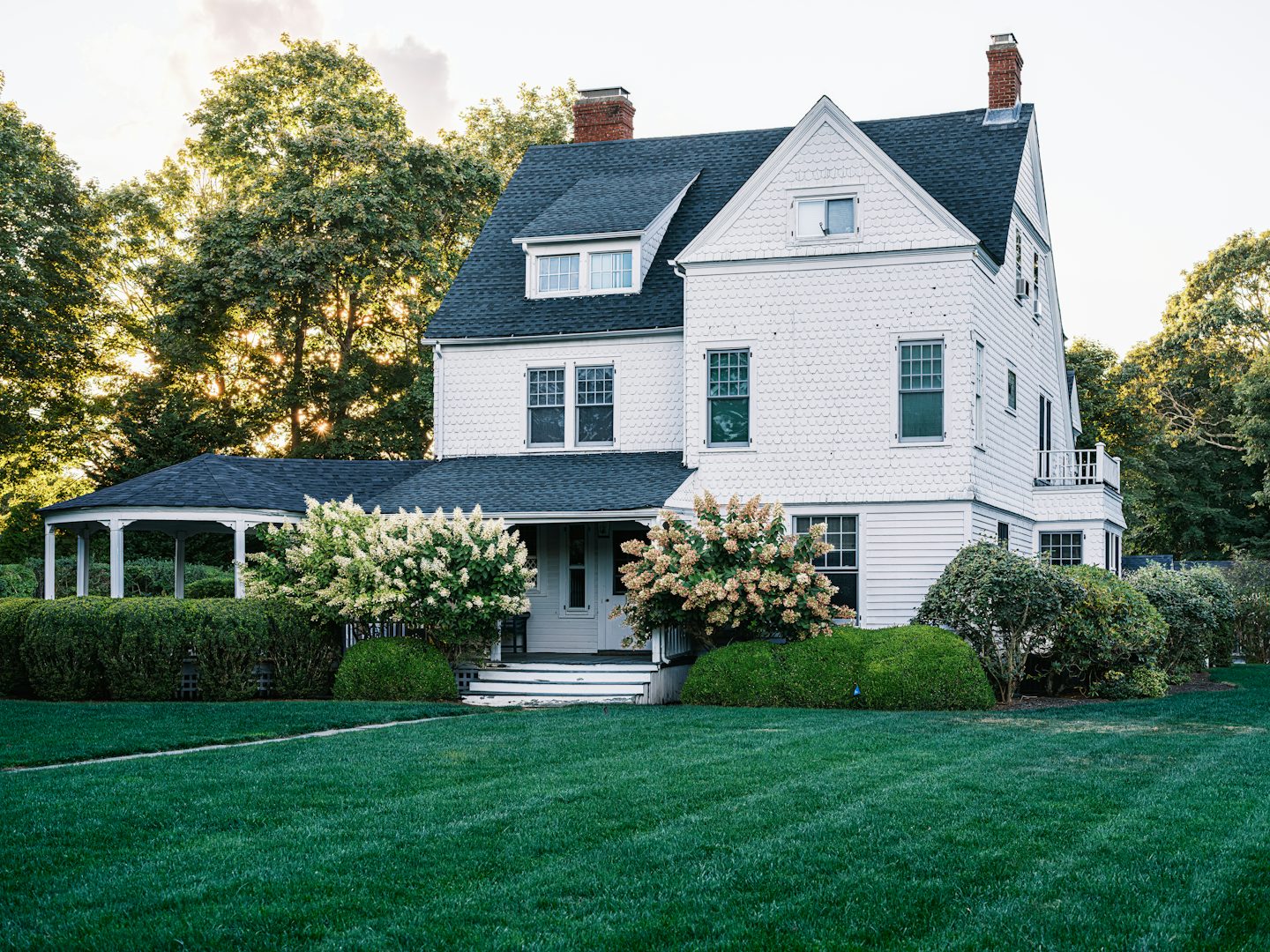 White shingled house with a wraparound porch and flowering hydrangeas on a wide lawn in late sun